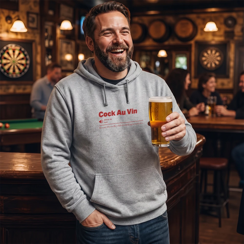 A smiling man in a bar wearing a light gray hoodie with red text that reads 'Cock Au Vin' and holding a glass of beer.
