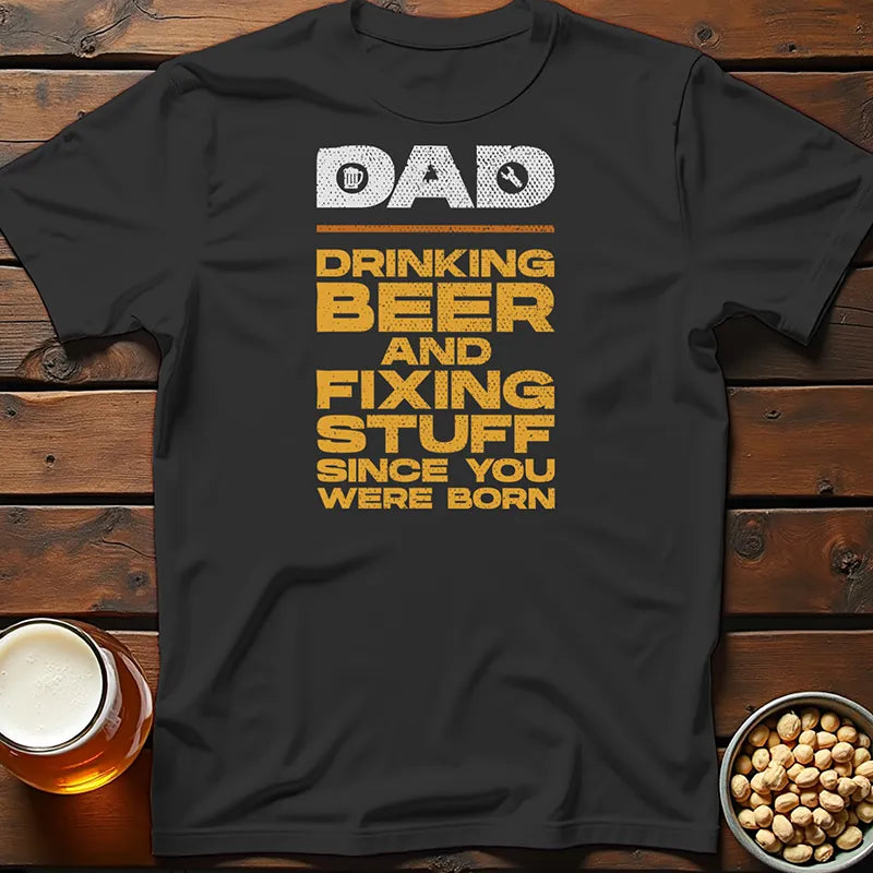 Black Father's Day t-shirt with yellow text on a wooden surface with a beer mug and bowl of nuts.