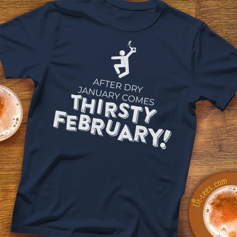 Navy blue t-shirt with 'After Dry January Comes Thirsty February!' text on a wooden surface with hot chocolate.