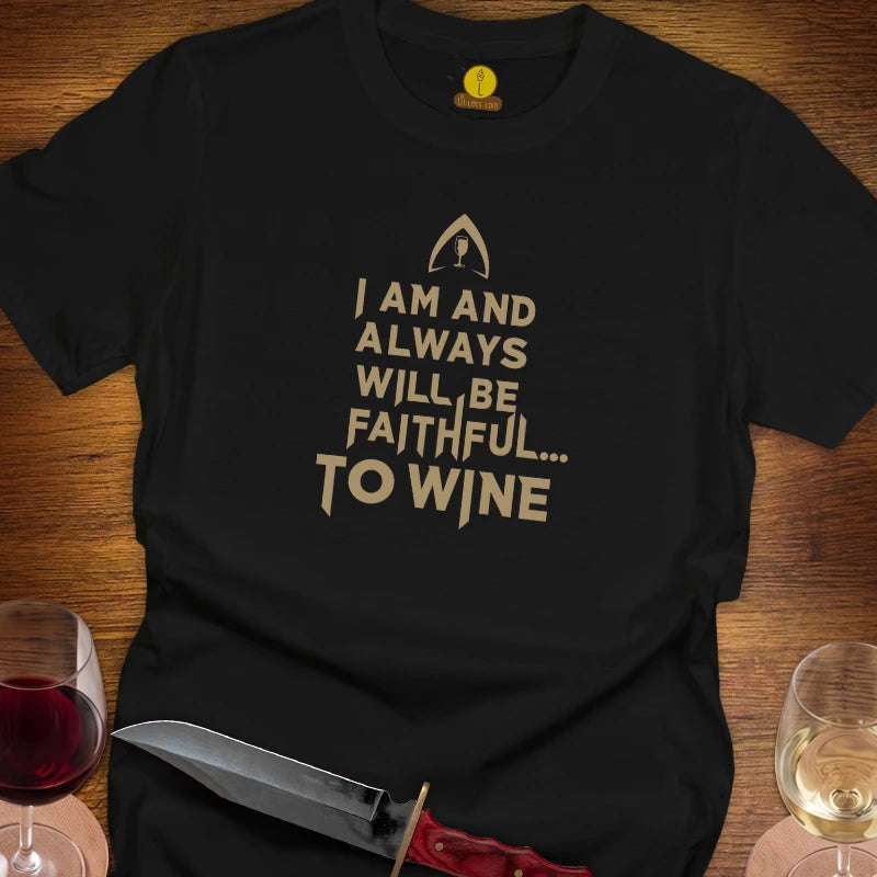 A black short-sleeve graphic T-shirt laid flat on a wooden surface, with a gold printed text design reading 'I AM AND ALWAYS WILL BE FAITHFUL TO WINE'.