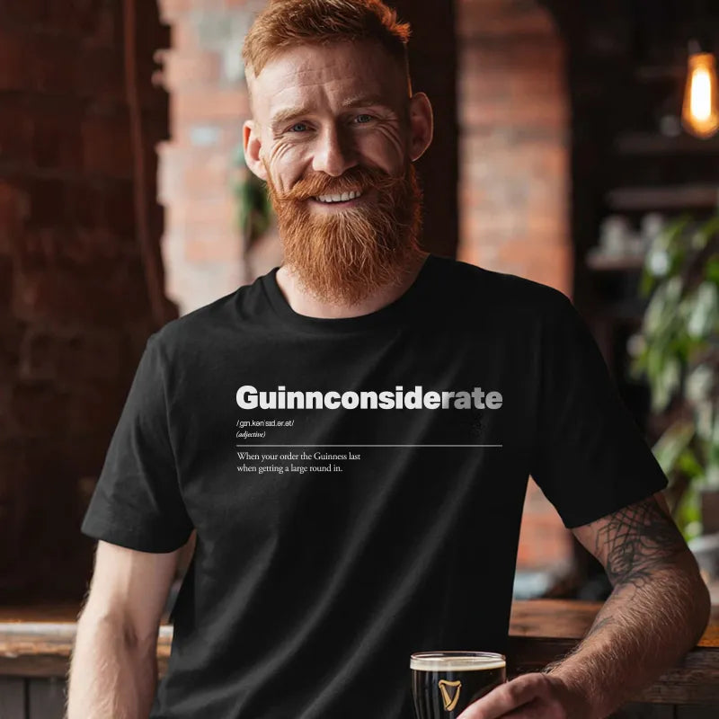 Man wearing a black t-shirt with text, holding a glass of beer in a pub setting. Guinnconsiderate :: When you order the Guinness last when getting a big round in.