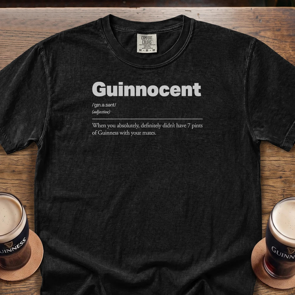 A black Comfort Colors 1717 Premium graphic Tee-shirt laid flat on a wooden table, featuring a white printed definition of the word 'Guinnocent' and reading: When you absolutely definitely didn't have 7 pints of Guinness with your mates. Two pint glasses of Guinness are partially visible at the bottom corners on wooden coasters.