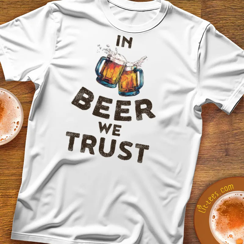 White t-shirt with 'In Beer We Trust' text and beer mug graphics on a wooden surface.