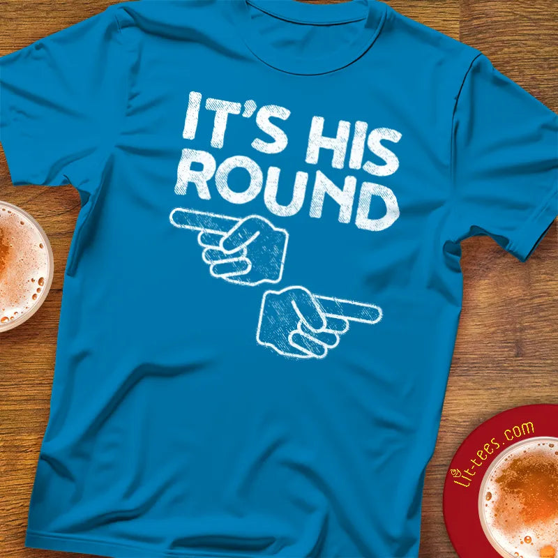 Blue t-shirt with 'It's His Round' text and pointing hands graphic on a wooden surface with beer glasses.