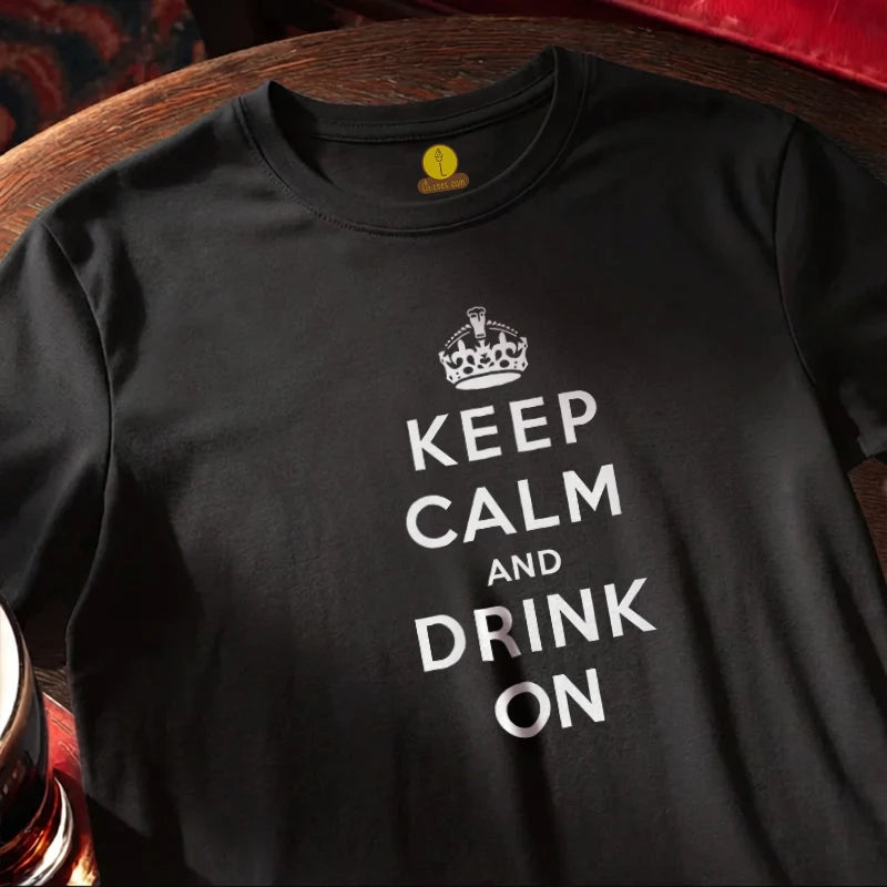 A black short-sleeve t-shirt with a white graphic print reading 'KEEP CALM AND DRINK ON' under a crown icon, laid flat on a wooden surface.
