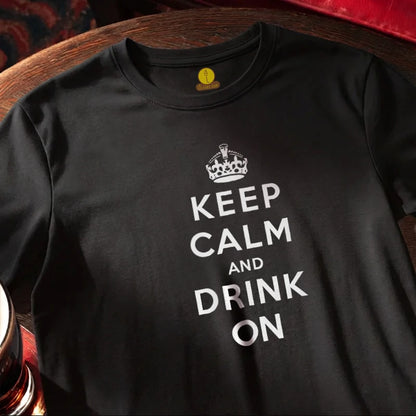 A black short-sleeve t-shirt with a white graphic print reading 'KEEP CALM AND DRINK ON' under a crown icon, laid flat on a wooden surface.
