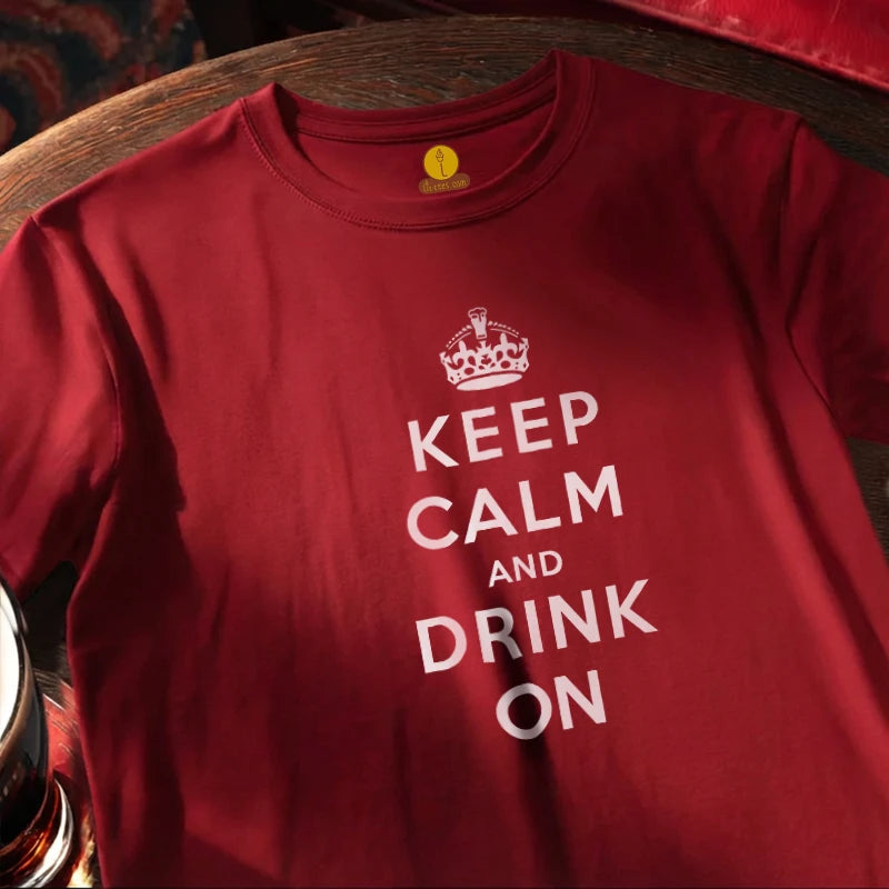 A red crewneck t-shirt featuring a white graphic print that reads 'KEEP CALM AND DRINK ON' with a crown icon above, laid flat on a dark wooden surface.