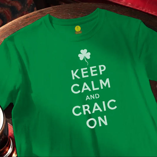 Green t-shirt with 'Keep Calm and Craic On' text and shamrock design on a wooden bar table.