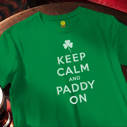 Green t-shirt with 'Keep Calm and Paddy On' text and shamrock design on a wooden bar table.