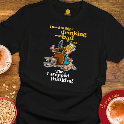 A black graphic t-shirt laid flat on a wooden surface, featuring a cartoon of a drunk man stumbling over a beer glass with the text 'I used to think drinking was bad for me. Then I stopped thinking.'