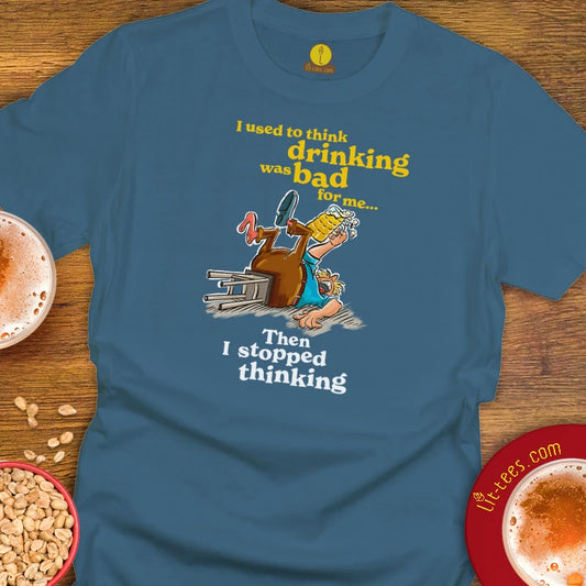 An Indigo graphic t-shirt laid flat on a wooden surface, featuring a cartoon of a drunk man stumbling over a beer glass with the text 'I used to think drinking was bad for me. Then I stopped thinking.'