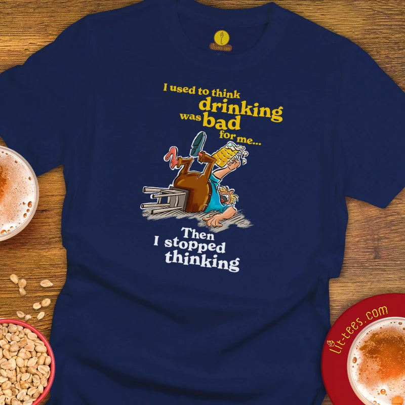 A Navy graphic t-shirt laid flat on a wooden surface, featuring a cartoon of a drunk man stumbling over a beer glass with the text 'I used to think drinking was bad for me. Then I stopped thinking.'