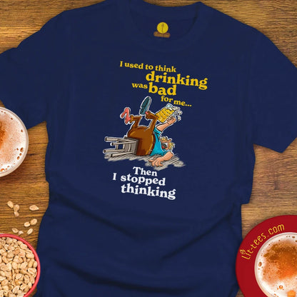 A Navy graphic t-shirt laid flat on a wooden surface, featuring a cartoon of a drunk man stumbling over a beer glass with the text 'I used to think drinking was bad for me. Then I stopped thinking.'