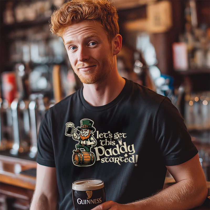 Man wearing a black t-shirt with a humorous design and text saying 'Let's Get This Paddy Started' holding a Guinness in a bar setting.