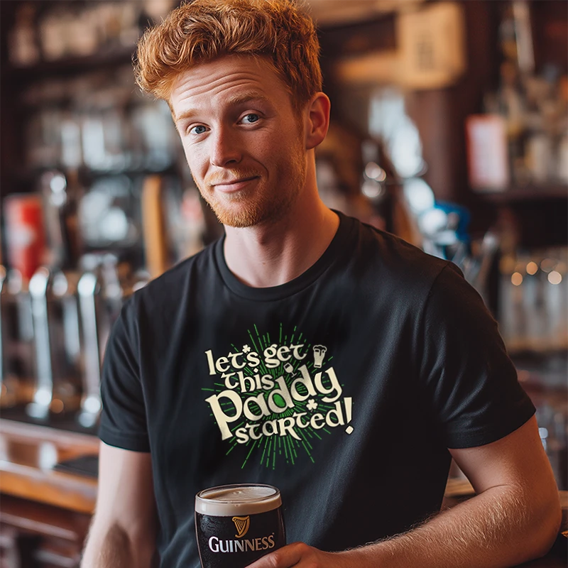 Man in a bar holding a pint of Guinness wearing a black t-shirt with text saying 'Let's Get this Paddy Started'