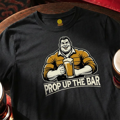 Black t-shirt with a graphic of a man holding a beer mug and 'Prop Up The Bar' text, placed on a wooden bar table.