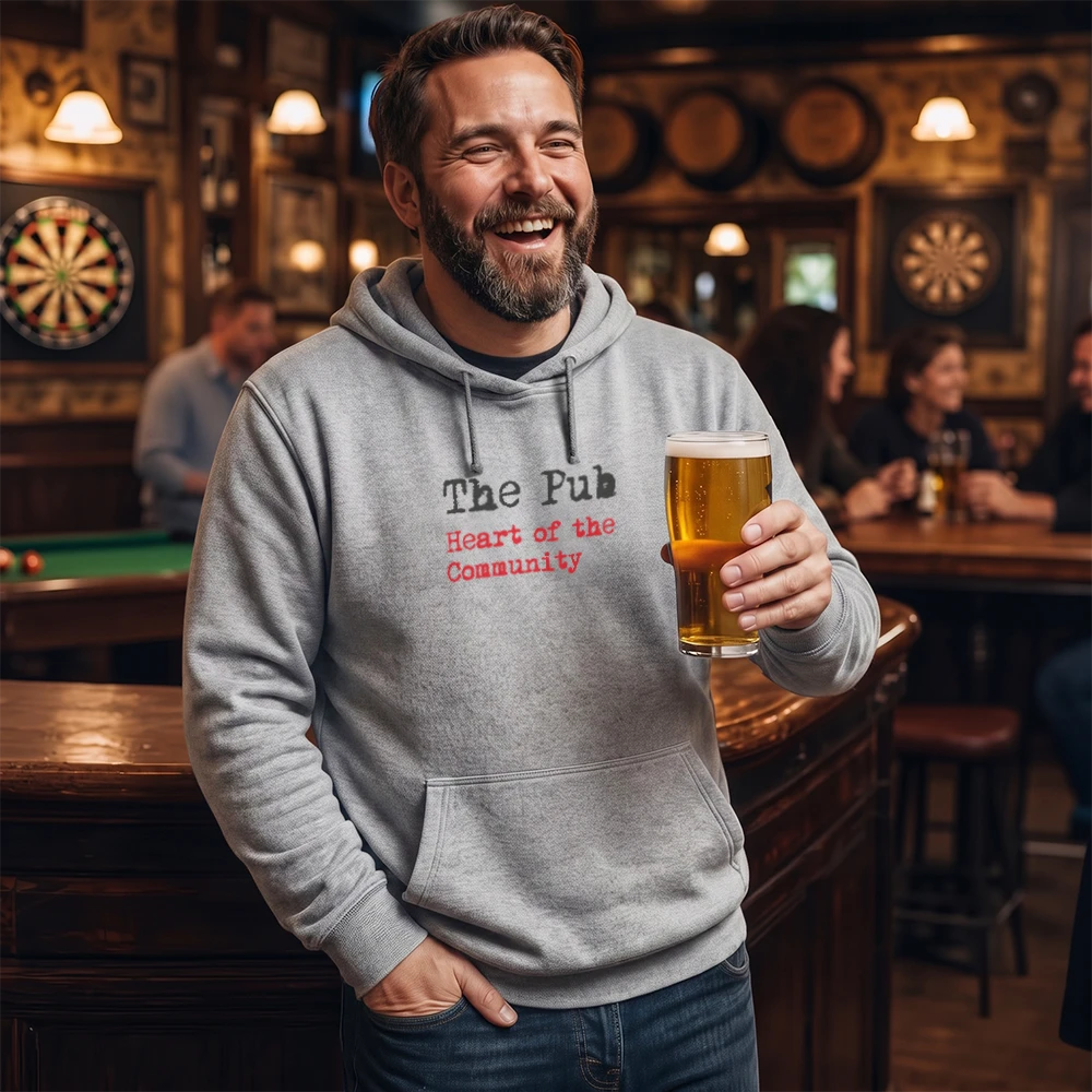 A smiling man in a bar wearing a light gray hoodie with red text that reads 'The Pub Heart of the Community' and holding a glass of beer.
