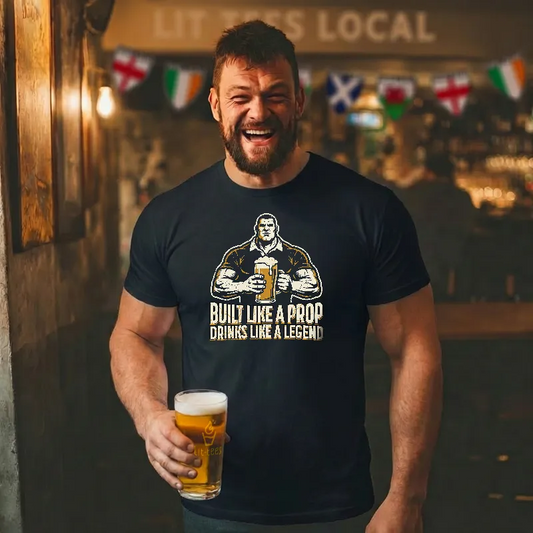 Man wearing a black t-shirt with text sayong 'Built like a prop drinks alike a legend' holding a beer in a bar setting.