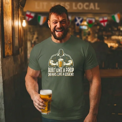 Man wearing a green t-shirt with a graphic design and text saying 'Built like a prop drinks like a legend' holding a beer in a bar setting.