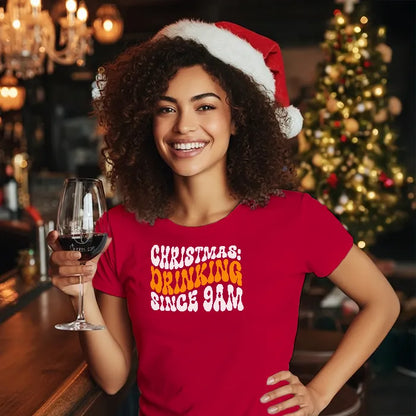 Lady in a pub setting wearing a Christmas themed tee shirt reading 'Christmas: Drinking since 9am' and holding a glass of wine. Glass has lit-tees.com printed on it.