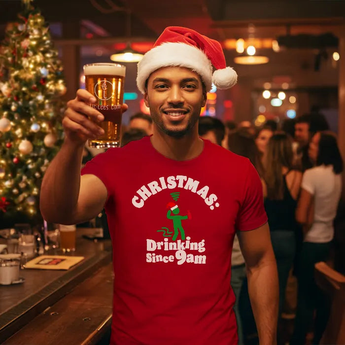 Man in a pub setting wearing a Christmas themed tee shirt reading 'Christmas: Drinking since 9am' and holding a glass of beer. Glass has lit-tees.com printed on it.