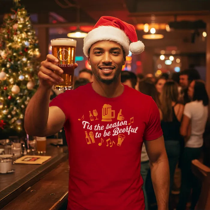 Man wearing a Santa hat and Christmas red shirt reading 'Tis the season to be beerful', holding a beer in a festive bar setting. Glass has lit-tees.com printed on it.