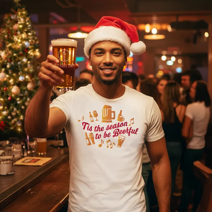 Man wearing a Santa hat and festive Christmas t-shirt reading 'Tis the season to be beerful', holding a beer in a bar setting with Christmas decorations. Glass has lit-tees.com printed on it.