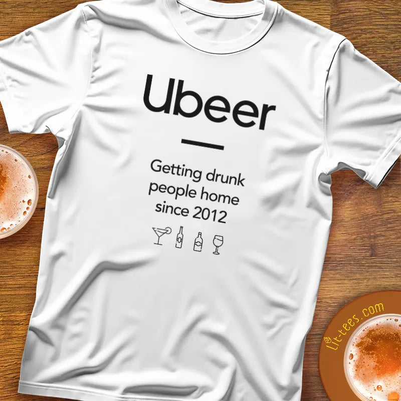 White t-shirt with 'Ubeer' text and graphics on a wooden surface with drinks.