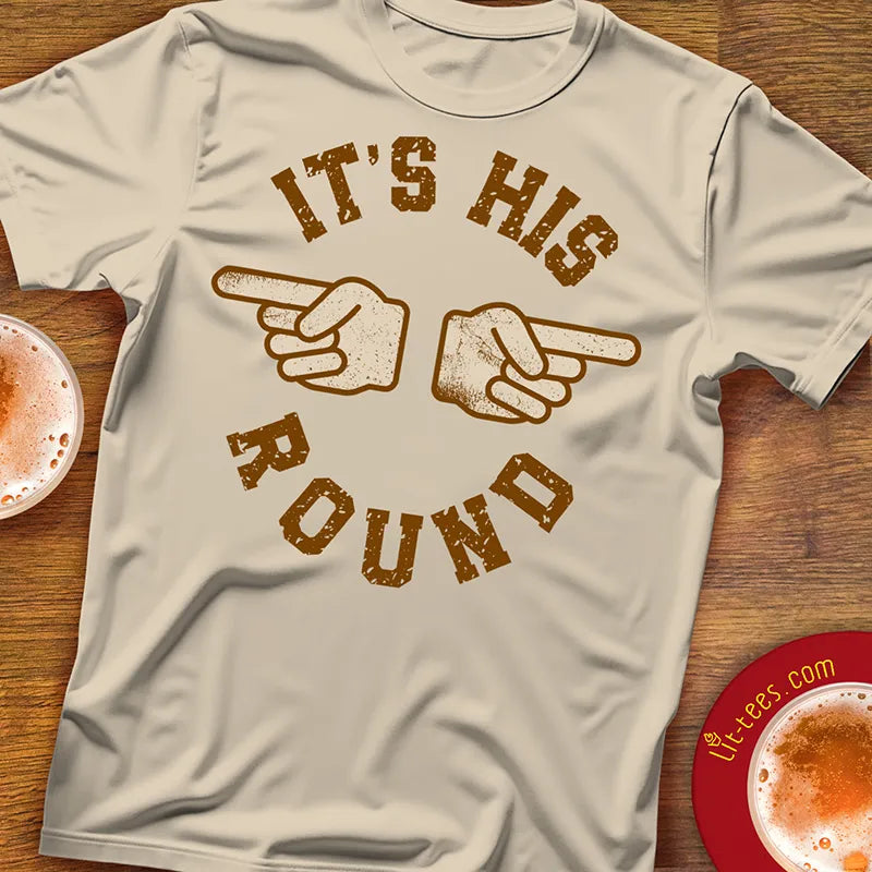 White t-shirt with brown graphic and text 'It's His Round' on a wooden surface with drinks.